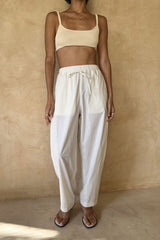 A model wearing loose white drawstring pants and a matching cream ribbed crop top, standing against a textured beige plaster wall.