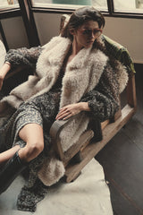Model wearing a fur vest in natural while sitting on a wooden chair indoors.