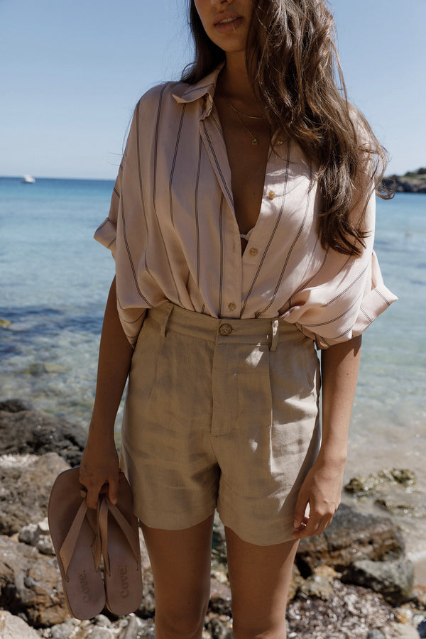 Woman in a light stripe blouse and beige shorts standing by the ocean